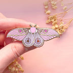 Pink butterfly-shaped moth pin held by a hand with a soft pink background
