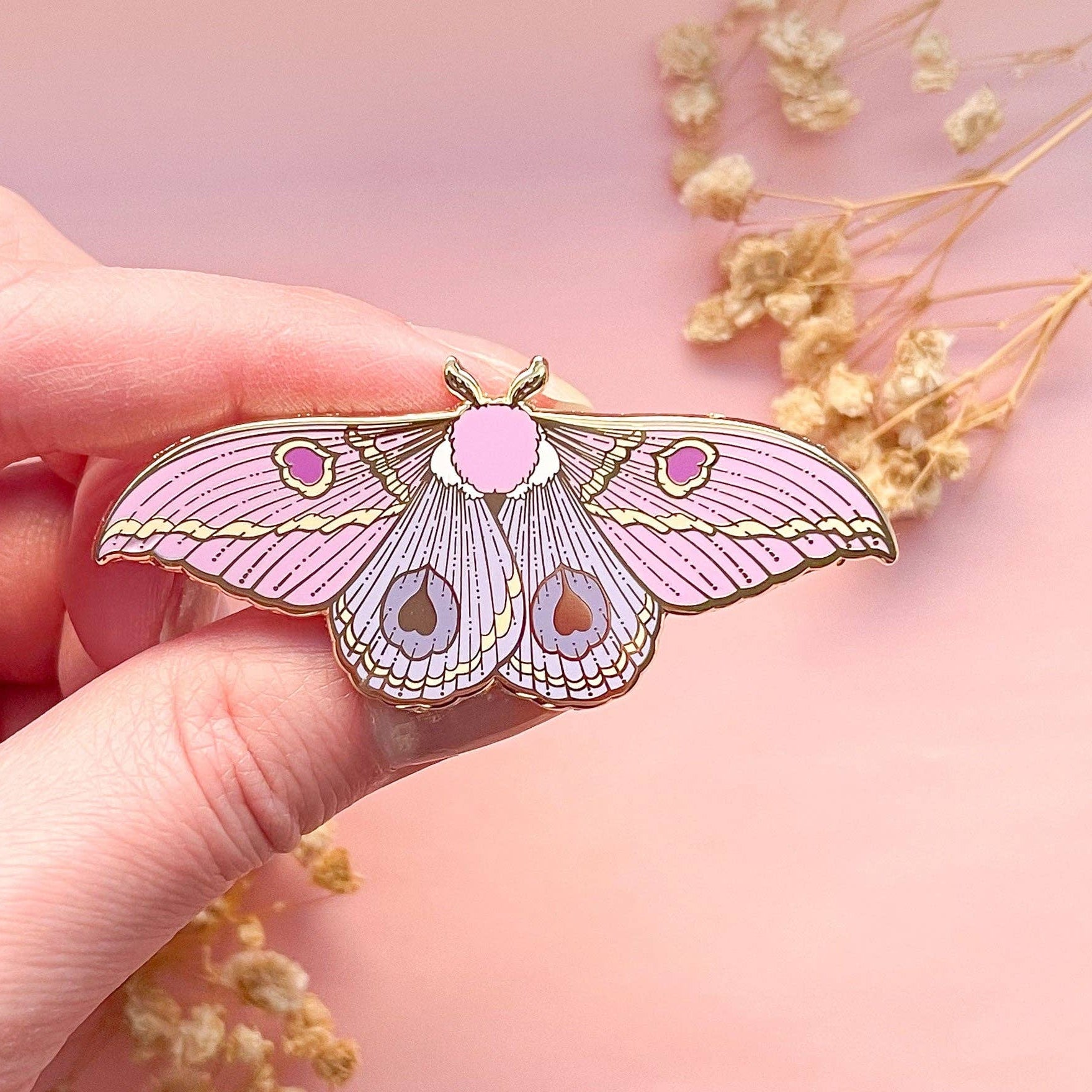 Pink butterfly-shaped moth pin held by a hand with a soft pink background