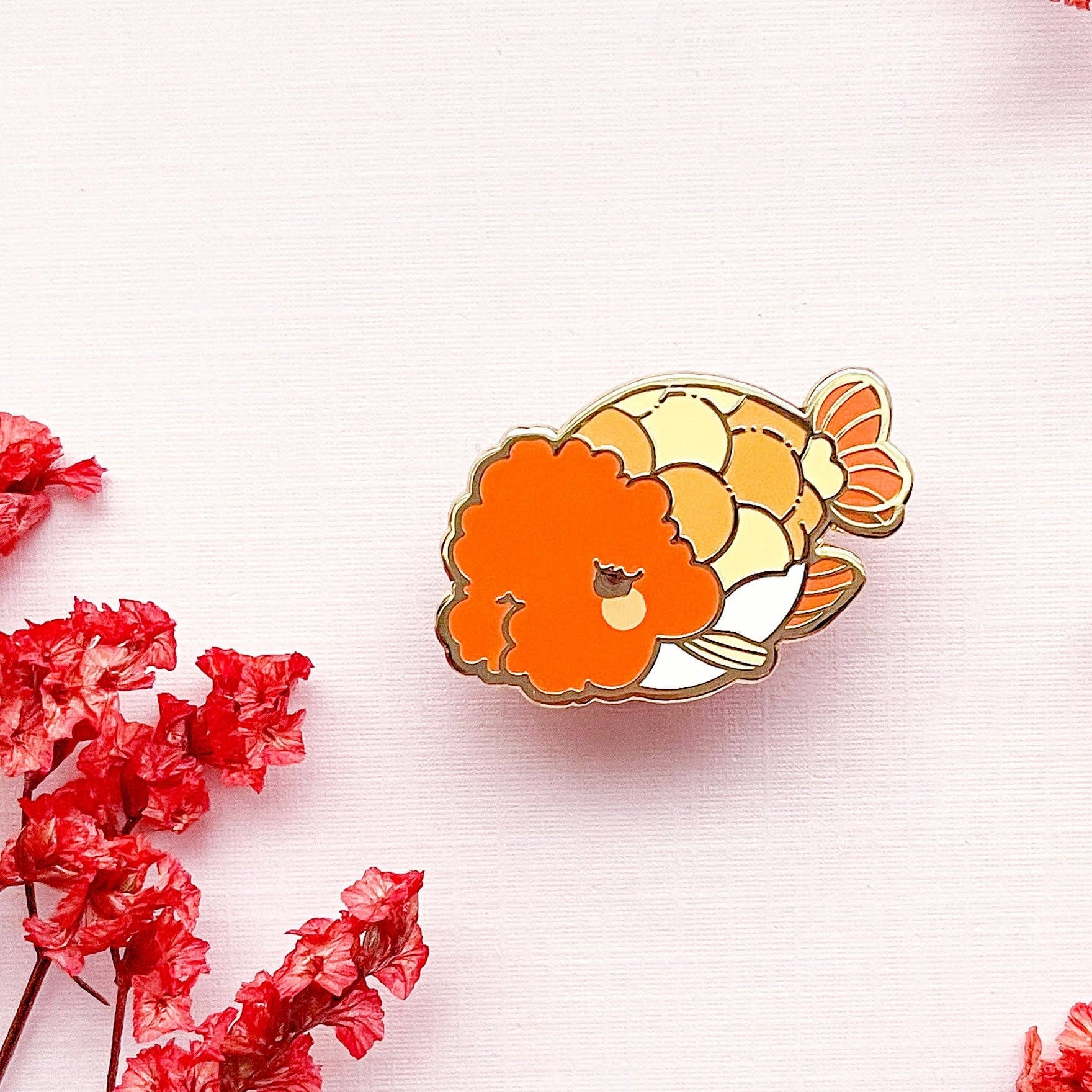 Colorful Ranchu Goldfish enamel pin on a background with red flowers.