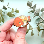 Hand holding a colorful Ranchu Goldfish enamel pin with green leaves in the background