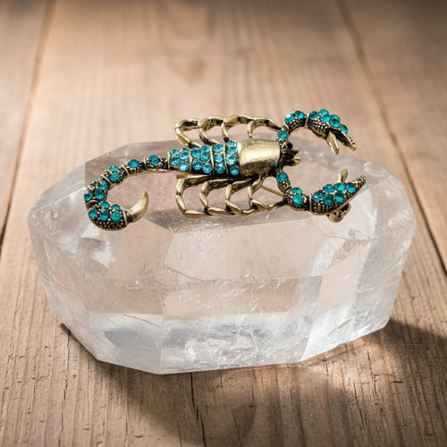 Scorpion-shaped brooch with turquoise stones on a wooden block, surrounded by green leaves.