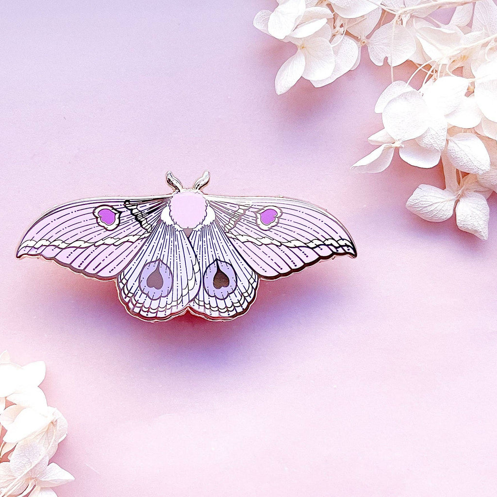 Butterfly-shaped moth pin with purple accents on a soft pink background with white flowers.
