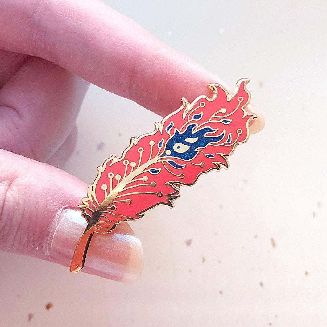 Feather-shaped phoenix pin with red, blue, and gold design held between fingers against a light background
