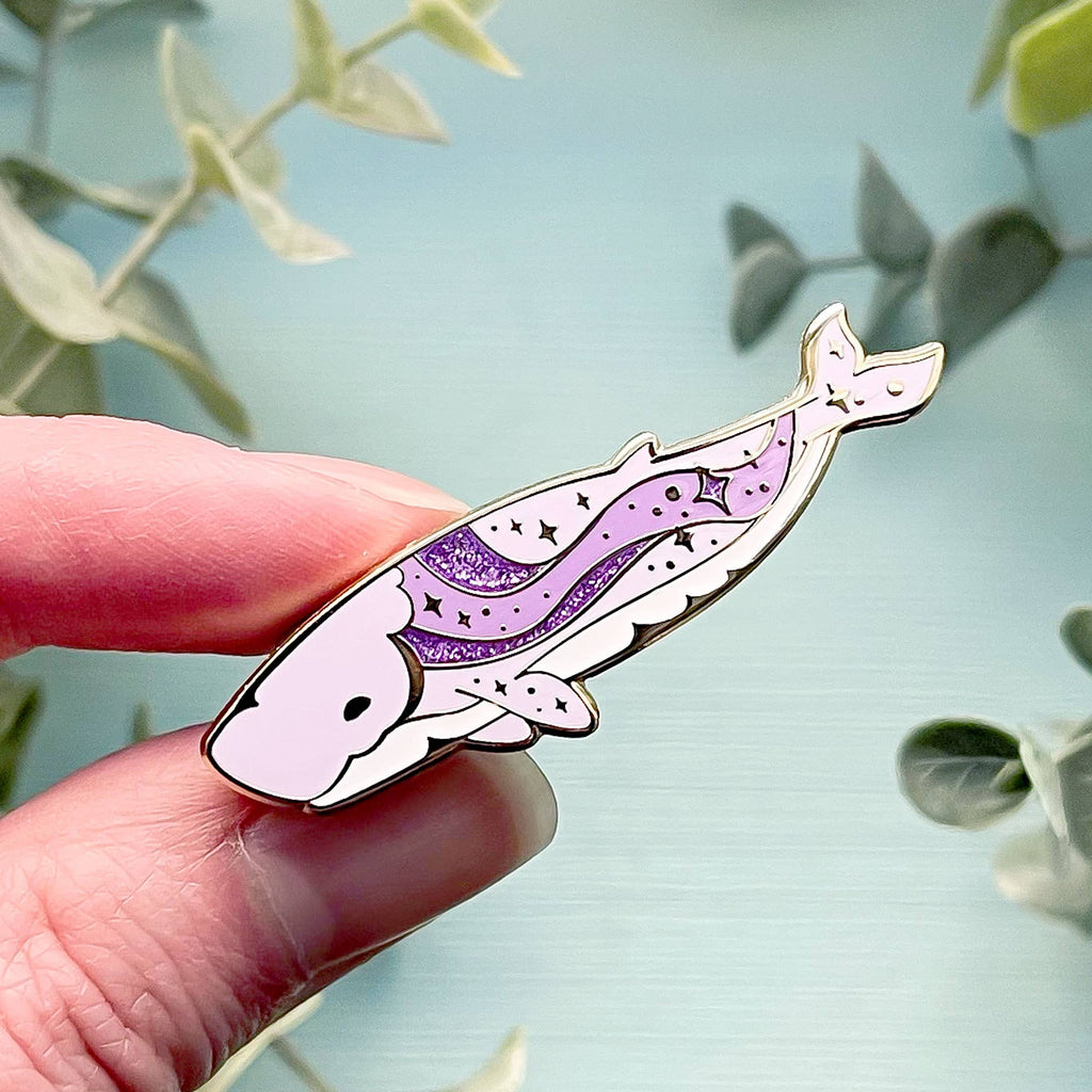 Hand holding a whale-shaped pin with purple and white design against a light blue background with green leaves.