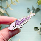 Hand holding a whale-shaped pin with purple and white design against a light blue background with green leaves.