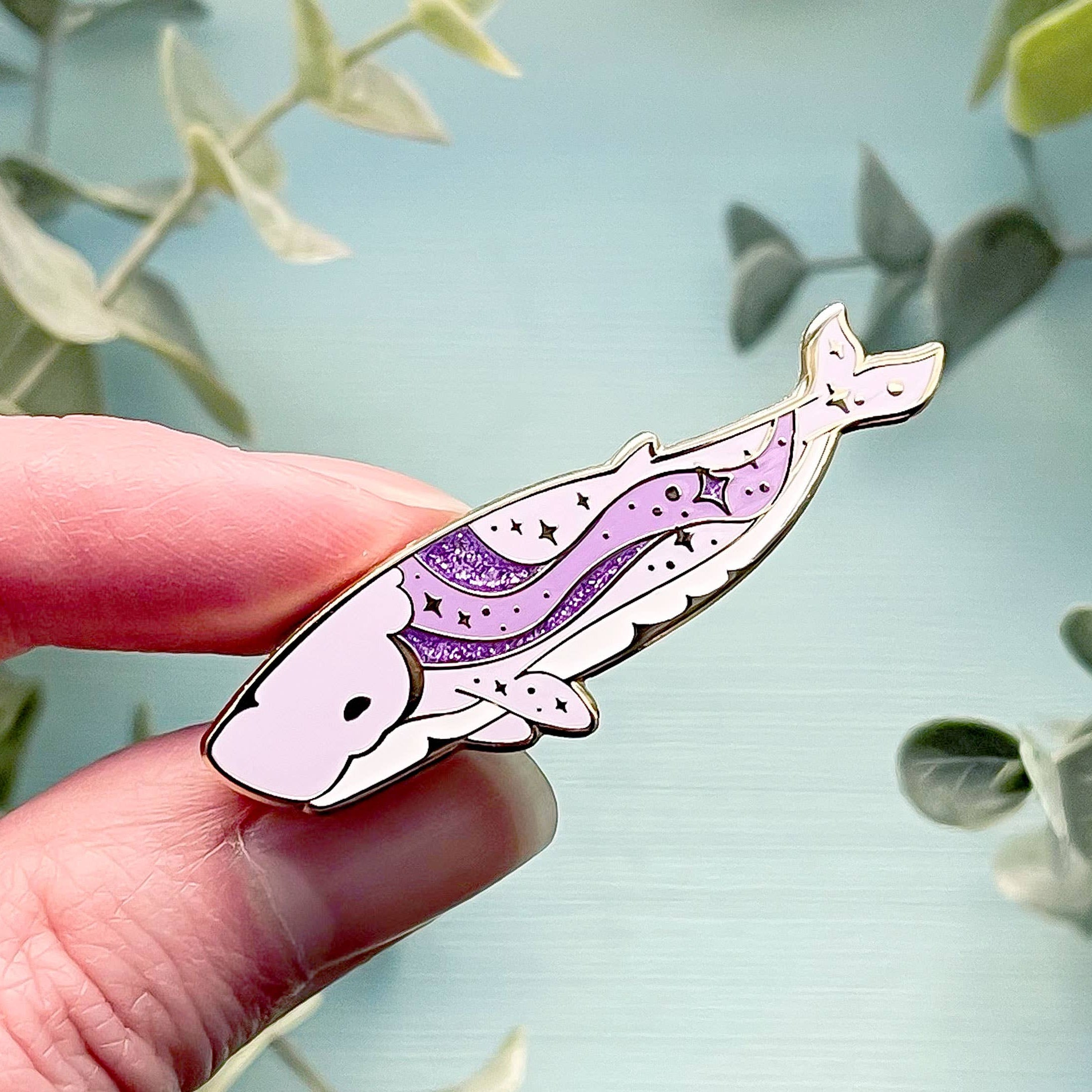 Hand holding a whale-shaped pin with purple and white design against a light blue background with green leaves.