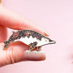 Dolphin-shaped brooch with gemstones held between fingers against a pink background