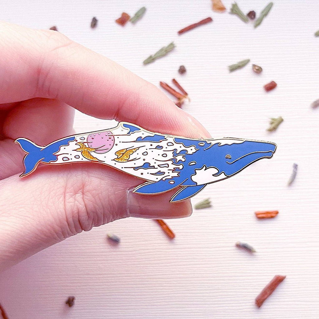 Hand holding a colorful whale-shaped pin against a light background with small decorative items.