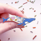 Hand holding a colorful whale-shaped pin against a light background with small decorative items.