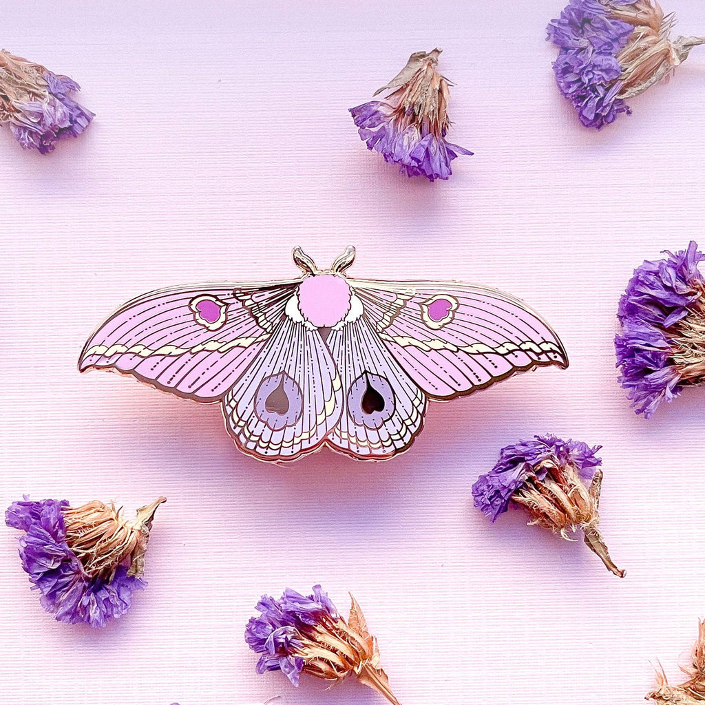 Moth-shaped enamel pin with pink and gold details on a light pink background with dried flowers.