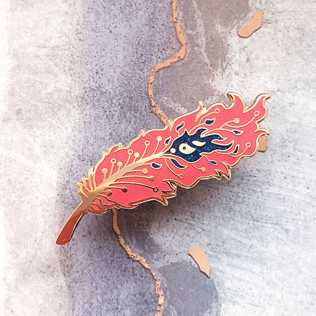 red feather-shaped pin with a blue eye design on a textured gray background
