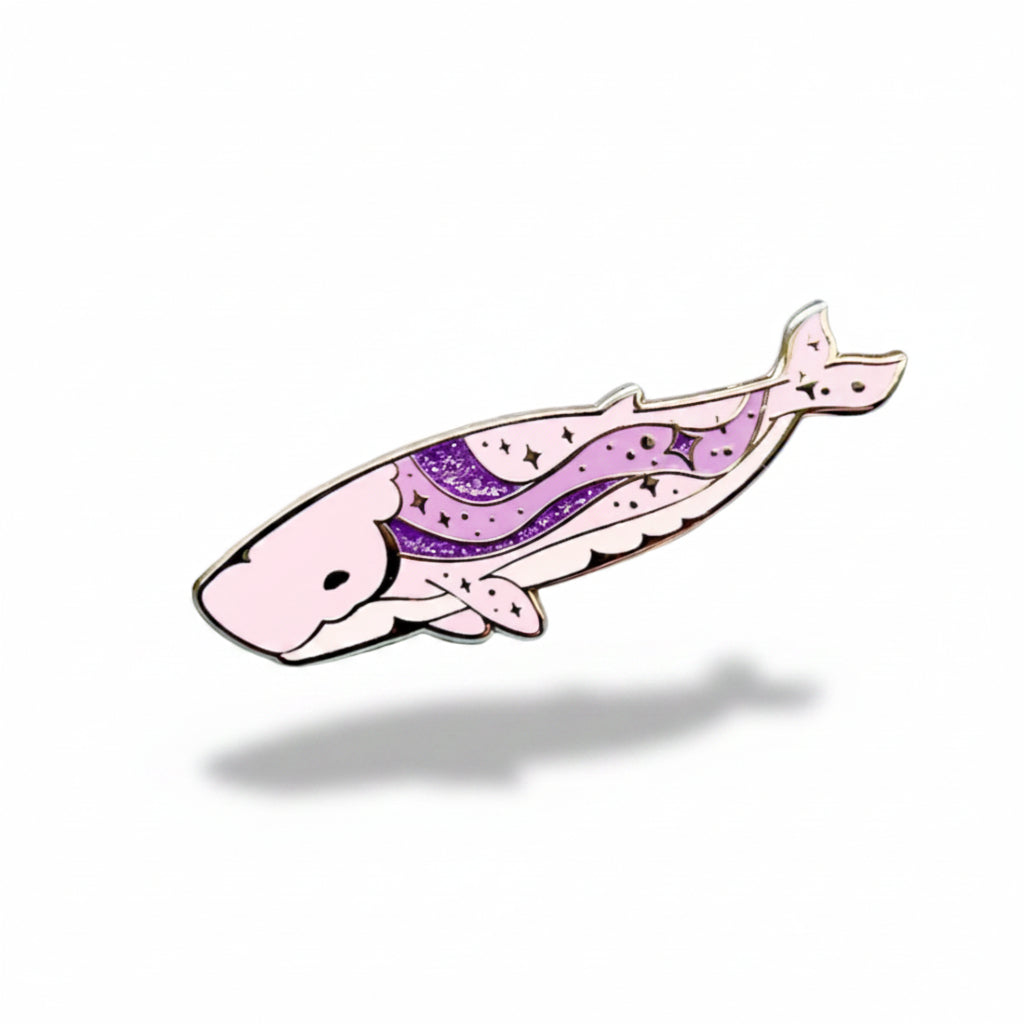 Pink and purple whale-shaped enamel pin on a light blue background with dried plants.