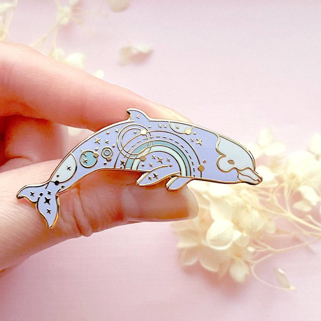 Hand holding a dolphin-shaped pin with a soft pink background