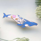 Decorative whale-shaped pin with a colorful design on a white background with greenery.