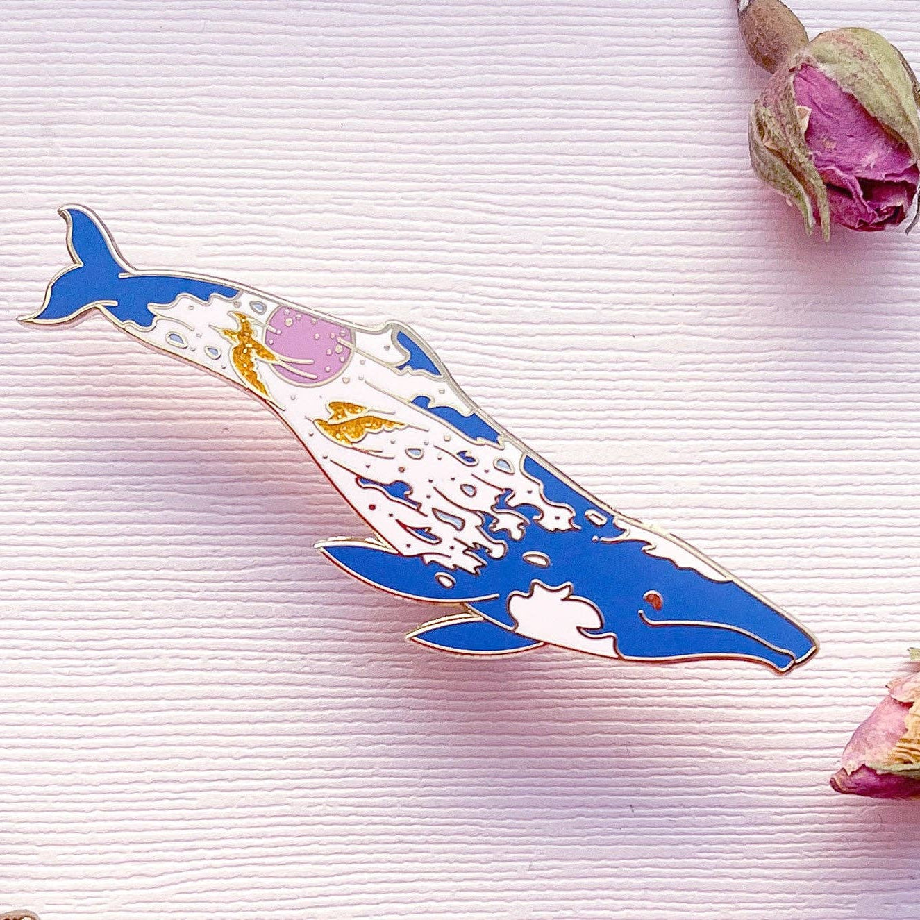 Colorful whale-shaped pin on a textured surface with dried flowers.