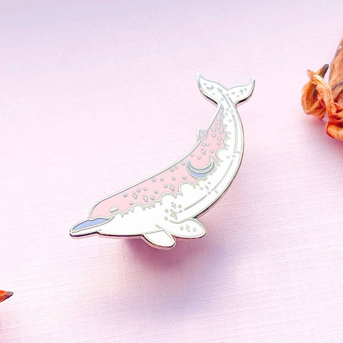 Decorative whale-shaped lapel pin on a light pink background with dried flowers.