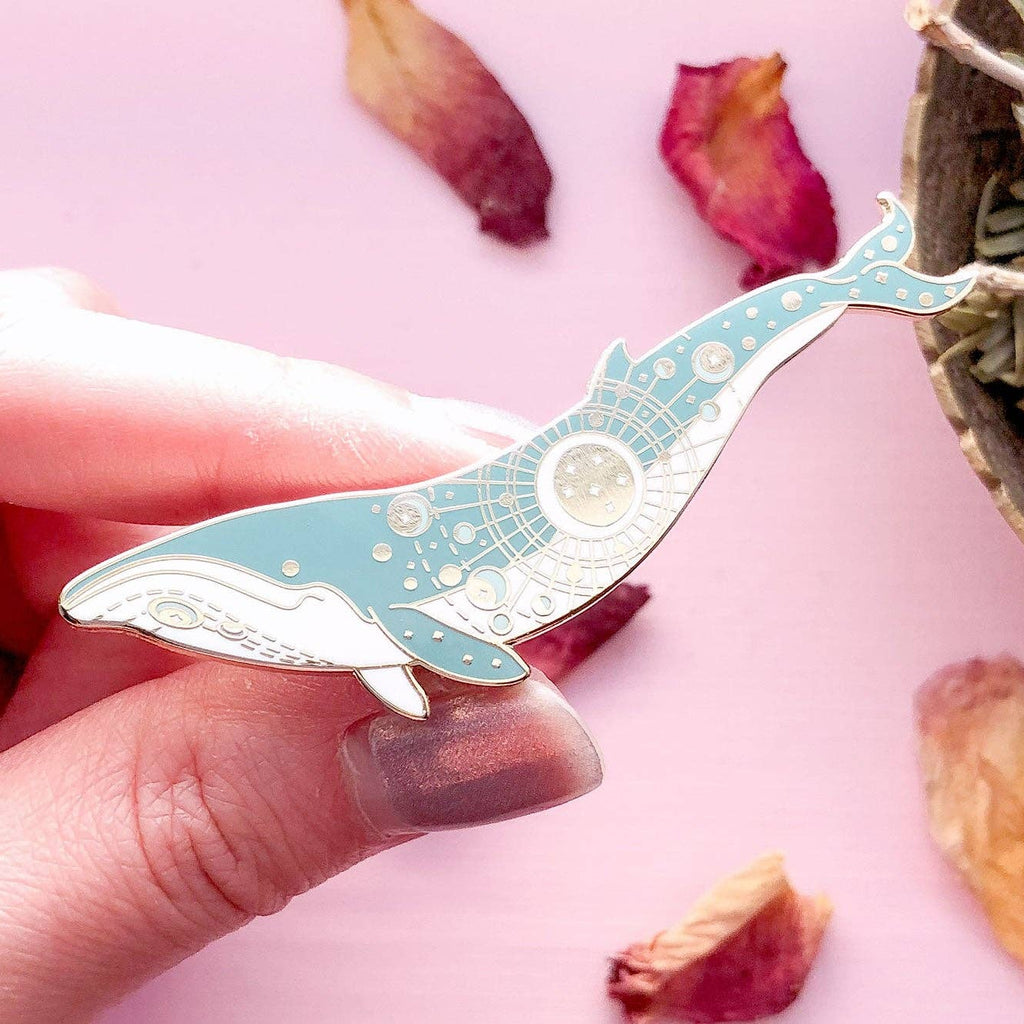 Hand holding a decorative whale-shaped item against a pink background with dried flowers and plants.
